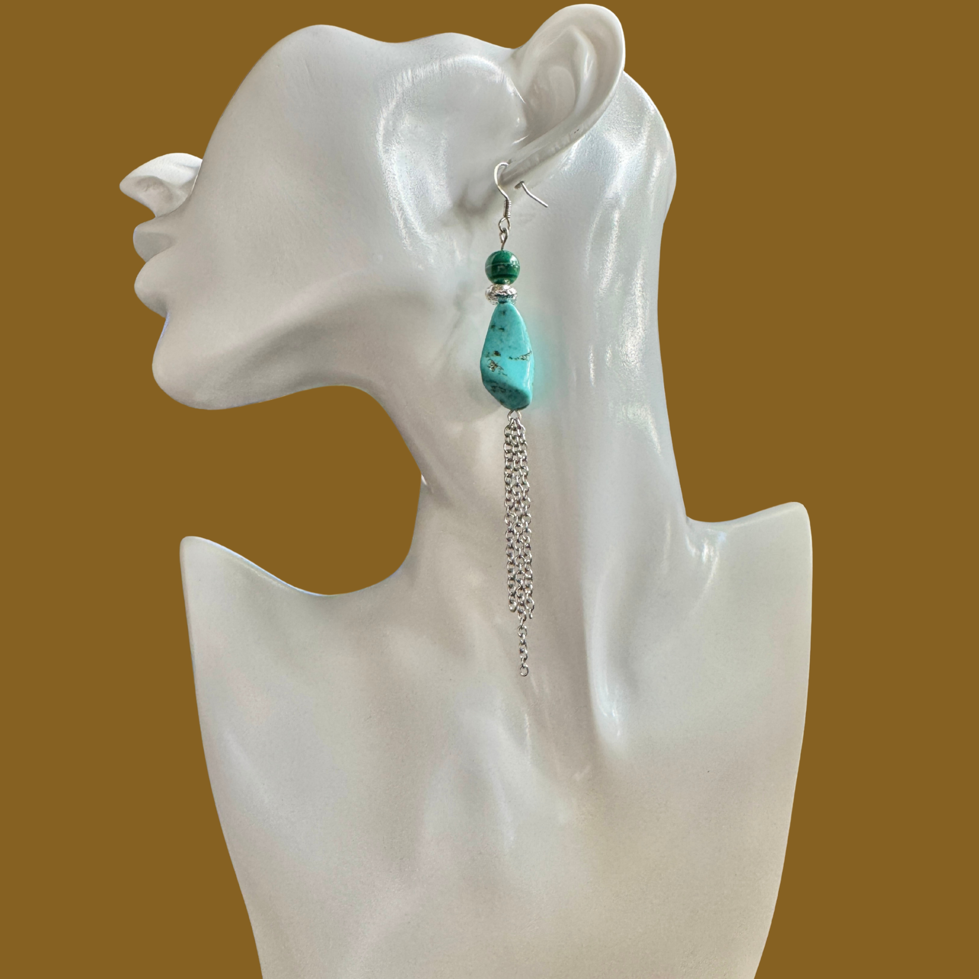 A single long dangle earring displayed on a white female mannequin head against a solid warm brown background. The earring features a turquoise-blue teardrop bead, a small green bead, a silver spacer ring, and a dangling silver chain.