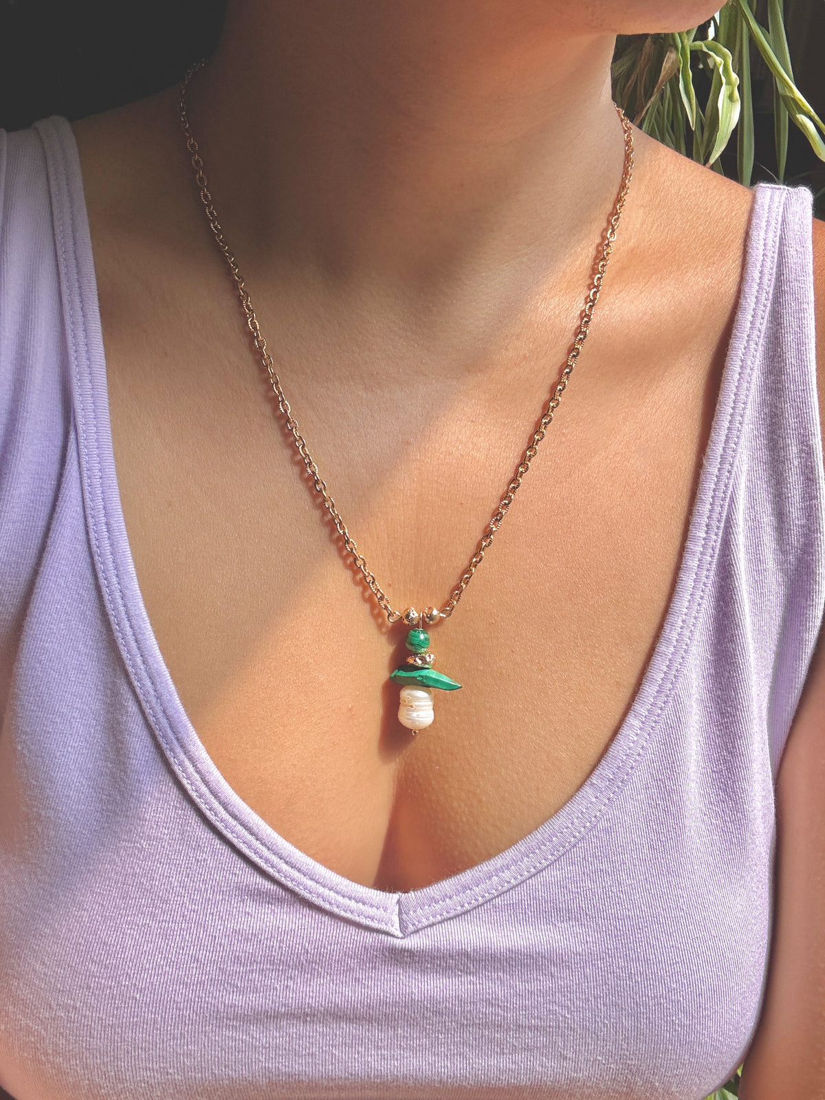 A close-up of a person wearing a delicate gold chain necklace with a small pendant featuring green and white details, styled with a light purple tank top.