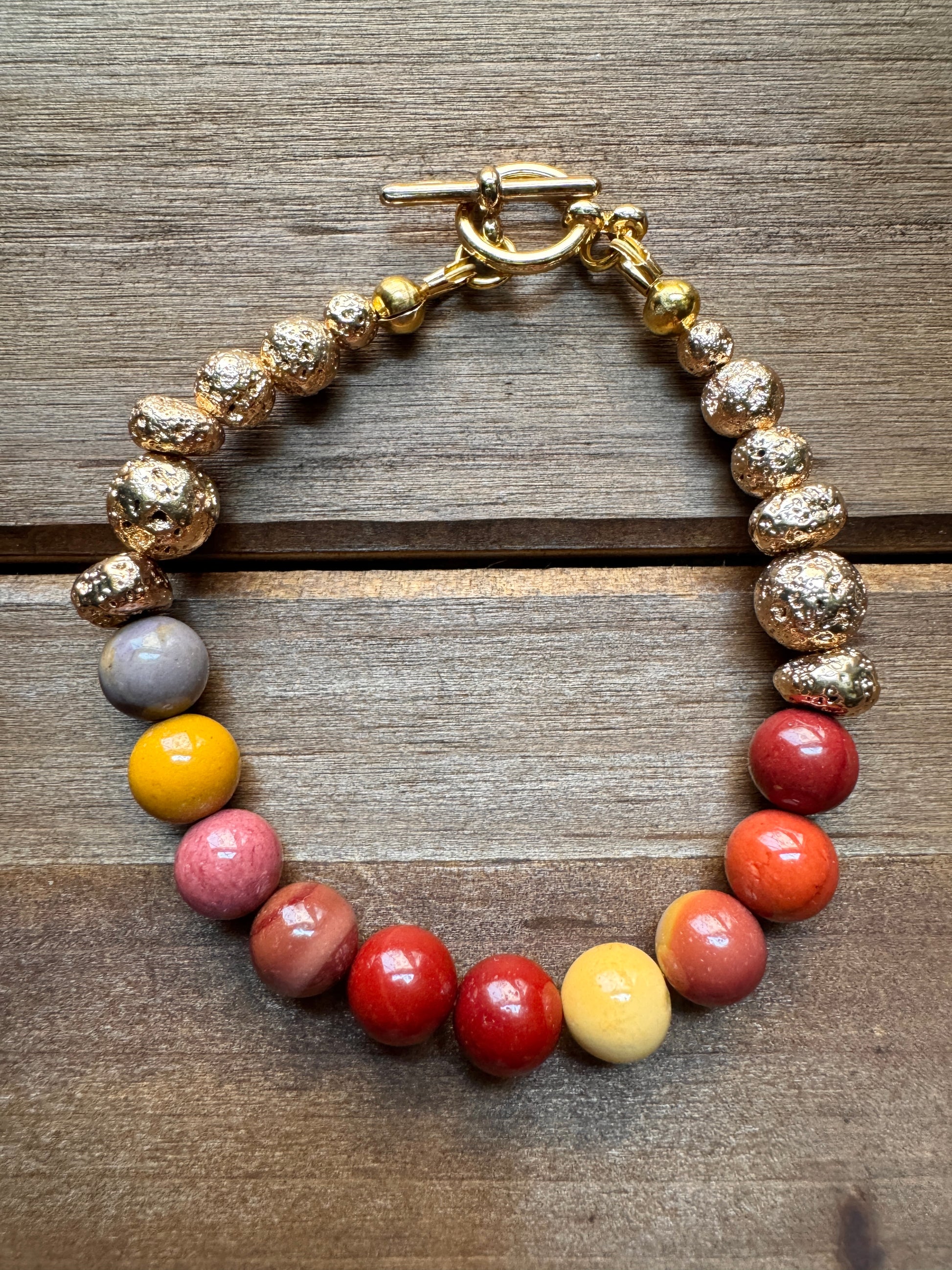 A beaded bracelet with textured gold-toned beads, a gray accent bead, and a row of glossy multicolor beads in yellow, pink, red, and orange, finished with a gold toggle clasp and photographed on a wooden surface.