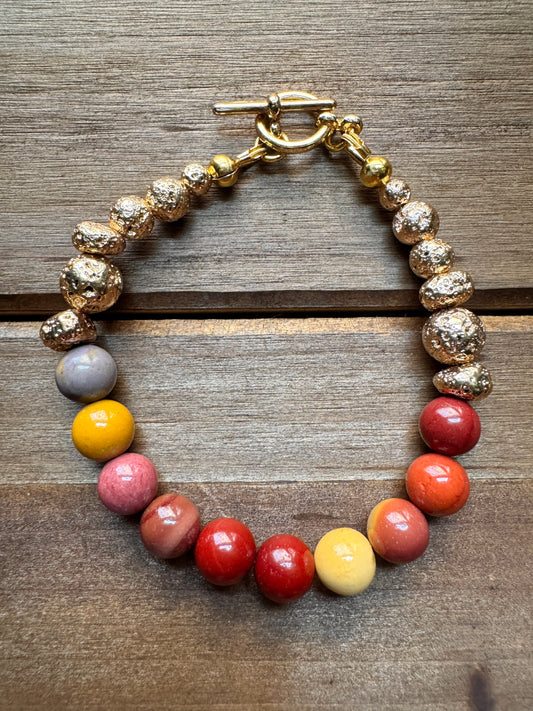 A beaded bracelet with textured gold-toned beads, a gray accent bead, and a row of glossy multicolor beads in yellow, pink, red, and orange, finished with a gold toggle clasp and photographed on a wooden surface.