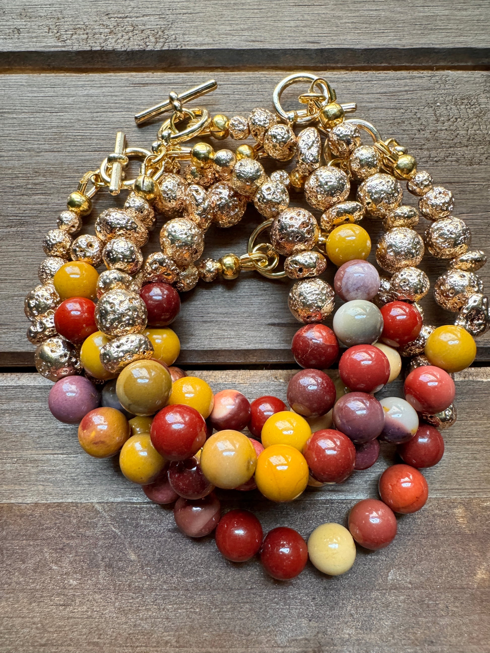 A multi-strand beaded necklace with gold-toned textured beads, yellow and red beads, and purple-gray beads, displayed on a rustic wooden surface.