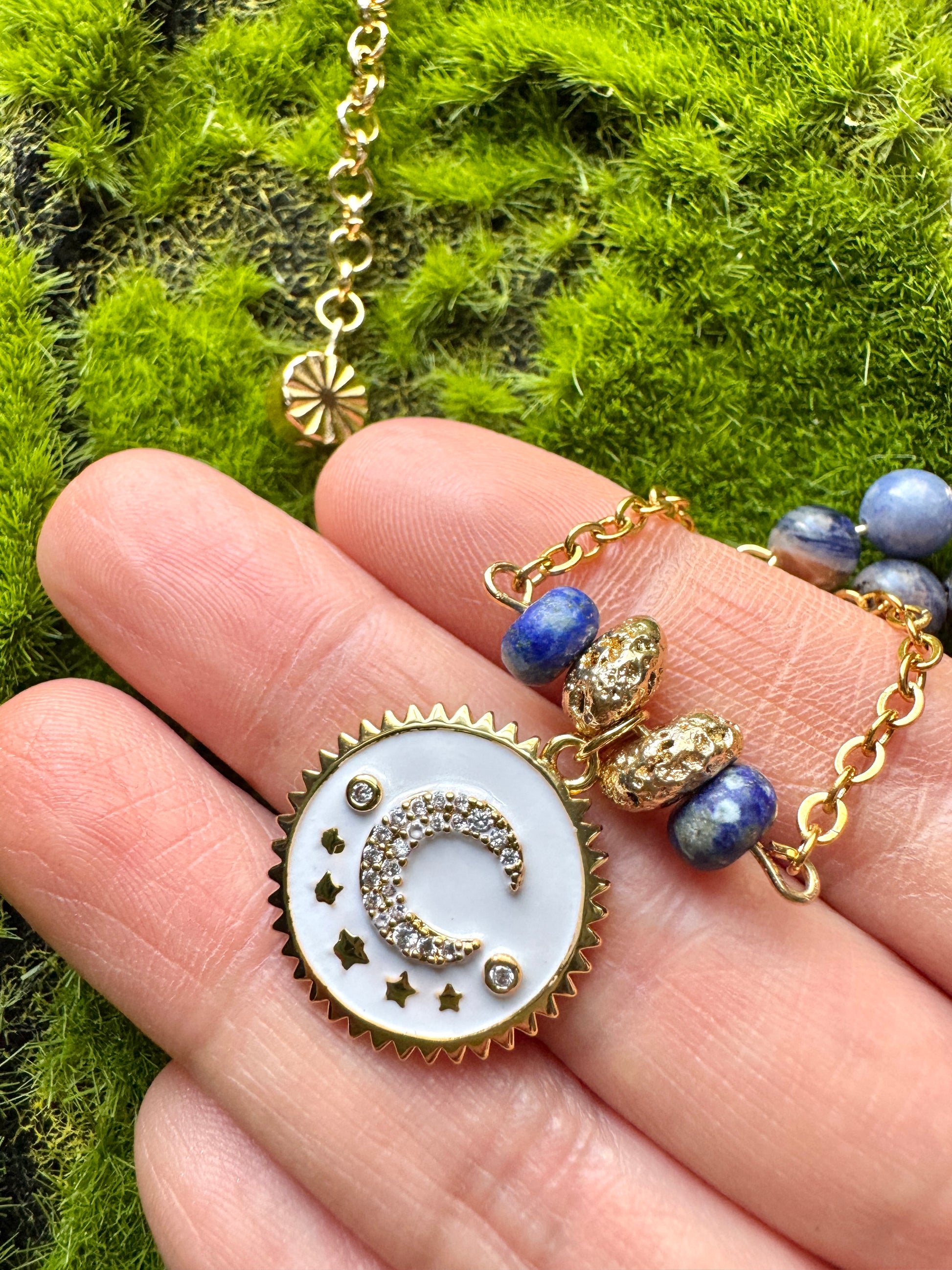 Gold necklace with a moon and star pendant held in a hand against a green moss background