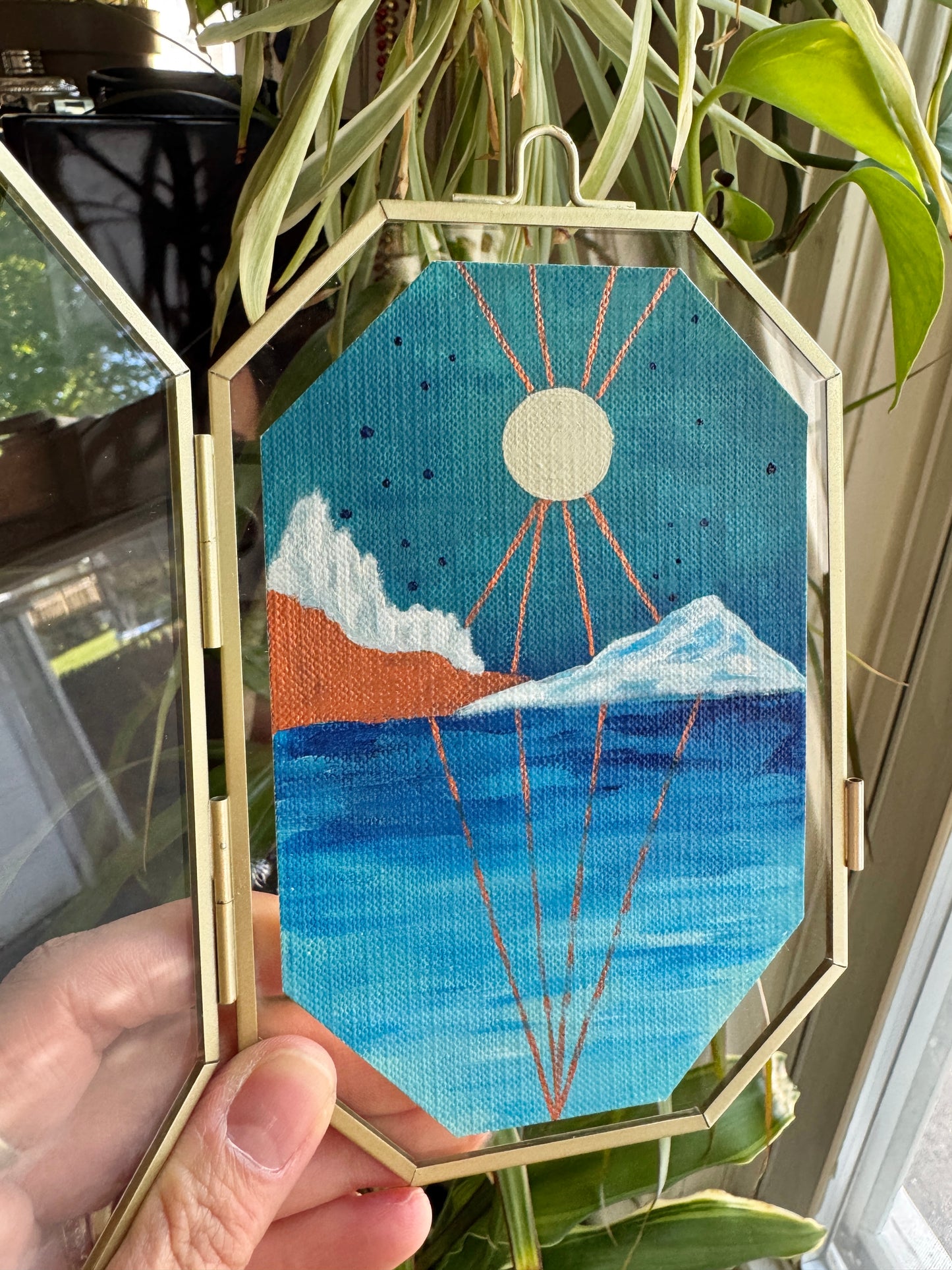 Hand holding up glass frame with landscape painting against a plant background