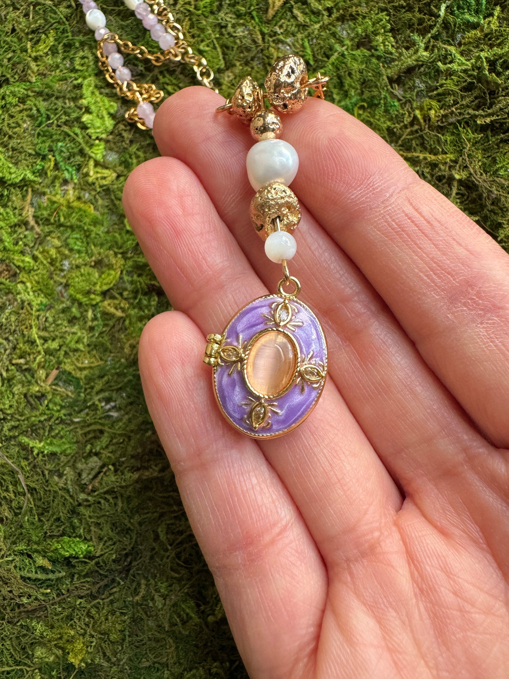 Necklace with a purple pendant held in a hand against a green moss background