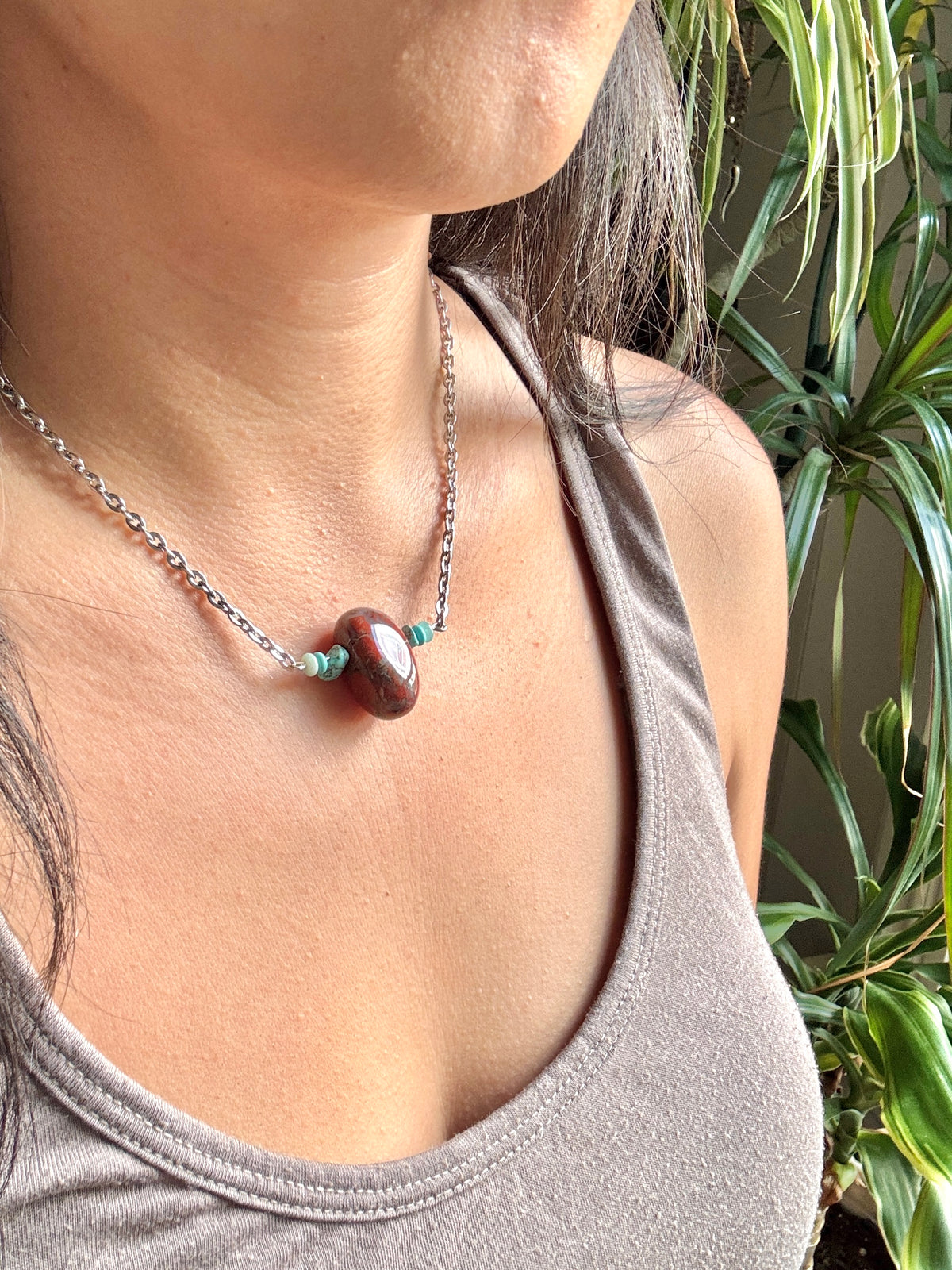 A close-up of a person wearing a silver chain necklace with a polished brown stone pendant and small turquoise spacer beads, paired with a gray tank top against a background of green houseplants.