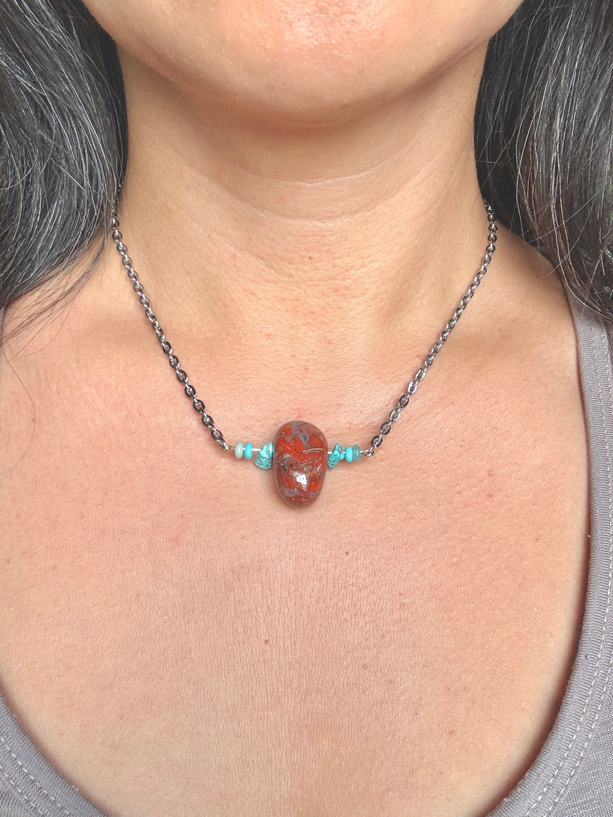 A close-up of a person's neck and chest wearing a dark metal chain necklace with a central oval red stone pendant and small turquoise-colored beads on each side.
