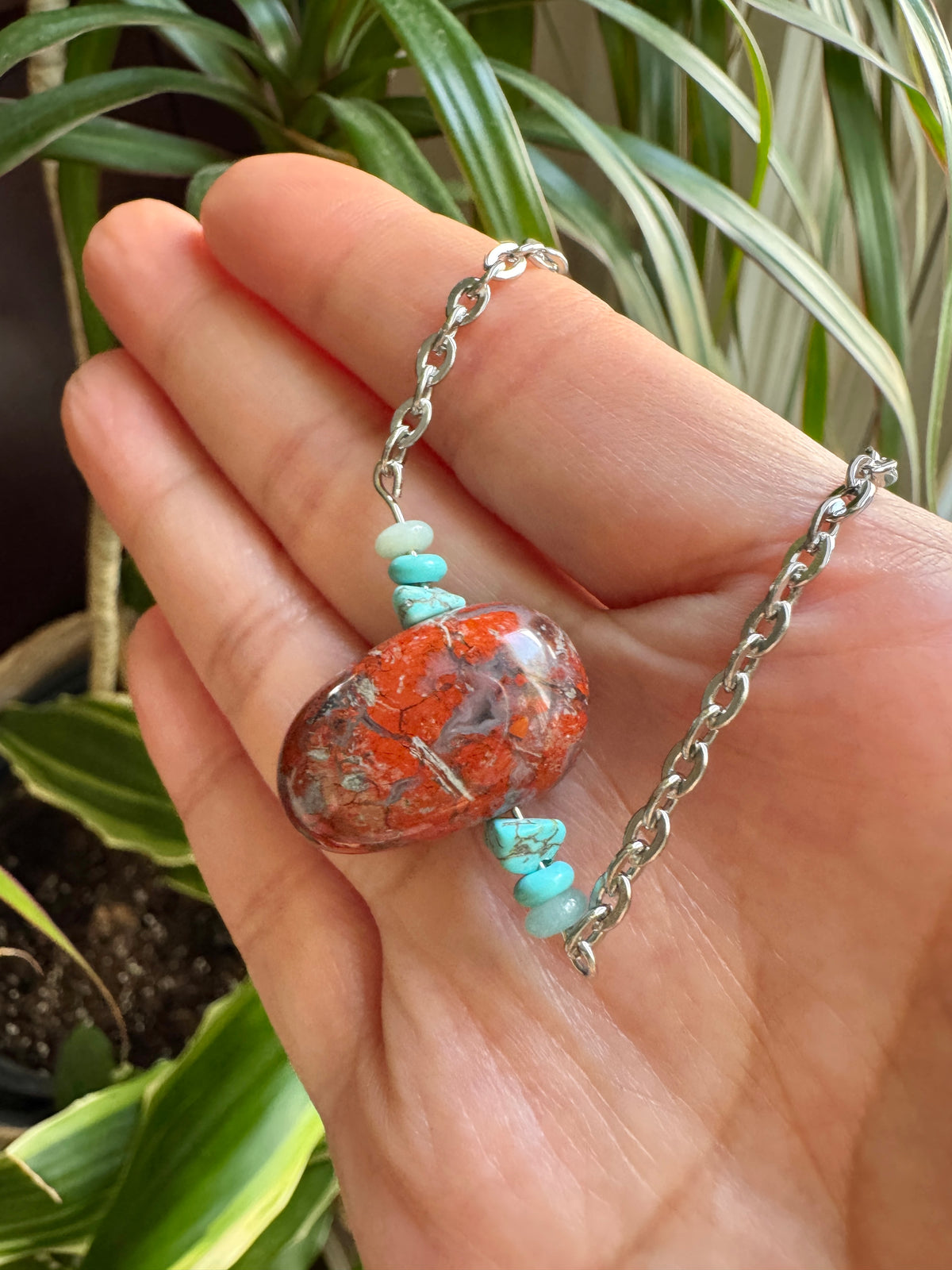 A silver chain necklace with a polished orange and white marbled stone pendant and small turquoise beads, held in a person's hand with green plants in the background.