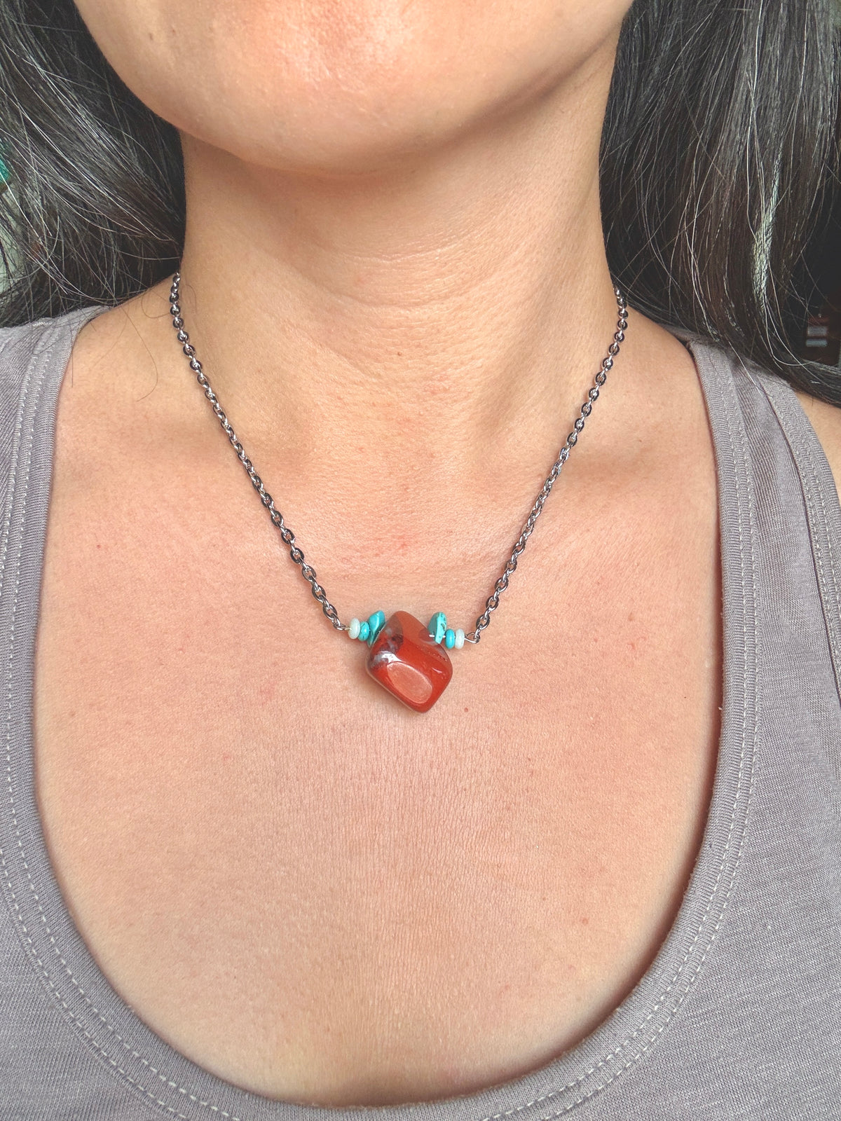 A close-up of a person's neck and chest wearing a silver chain necklace with a polished red-orange heart-shaped pendant and small turquoise accents.