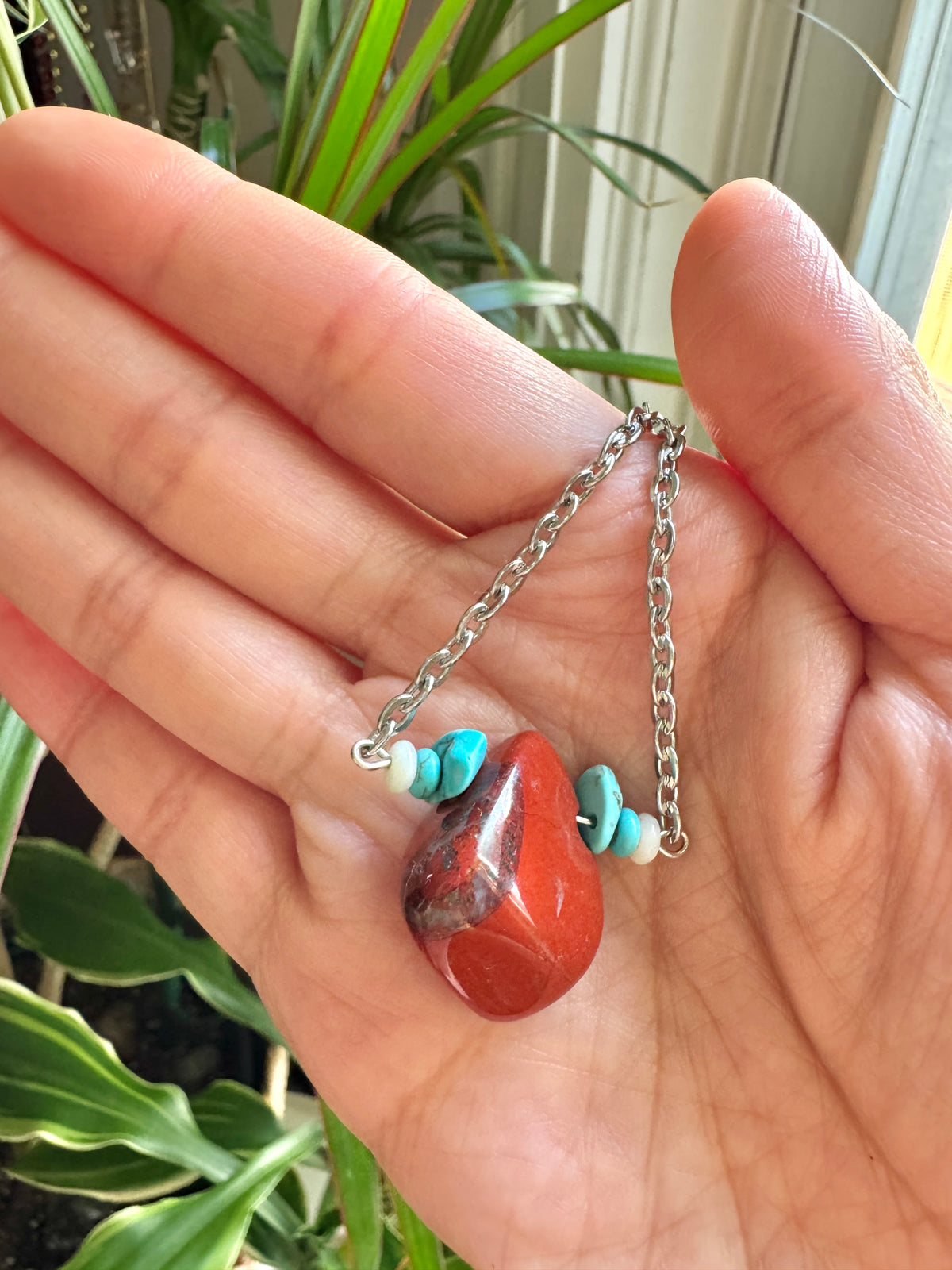 A close-up of a hand holding a silver chain necklace with a polished red-orange stone pendant and small turquoise beads, with green plants in the background.
