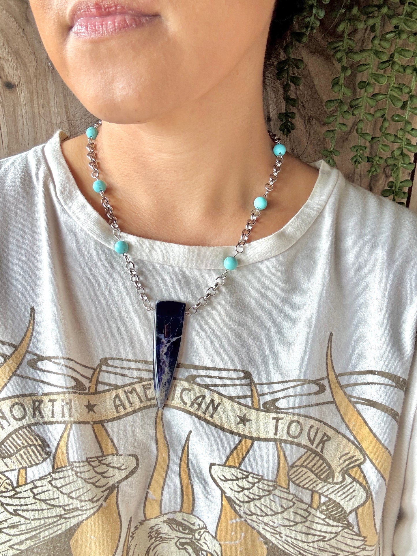 Person wearing a necklace with a blue pendant on a white shirt with gold text and graphics.