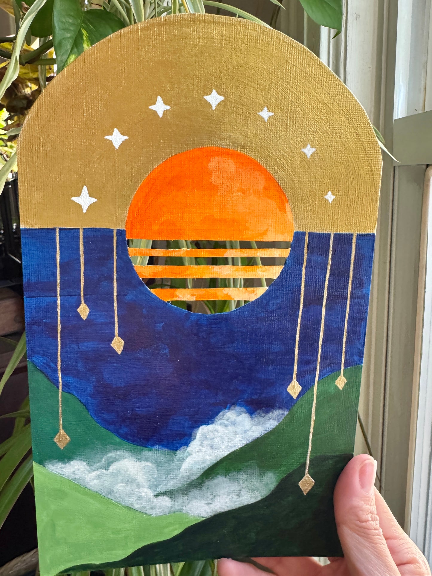 Hand holding up a landscape painting on paper against a plant background