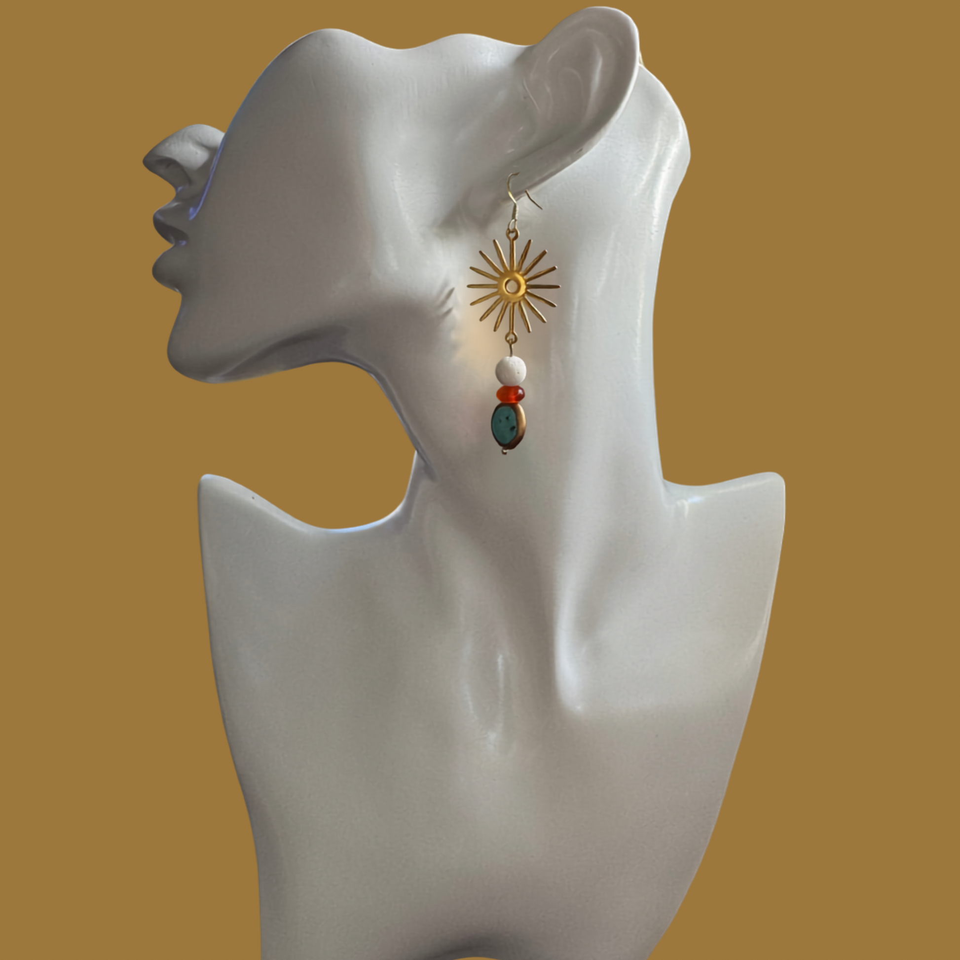 A single dangling earring displayed on a white female bust against a warm tan background. The earring has a gold sunburst top with a small white bead, a red spacer bead, and a dark teal oval stone.