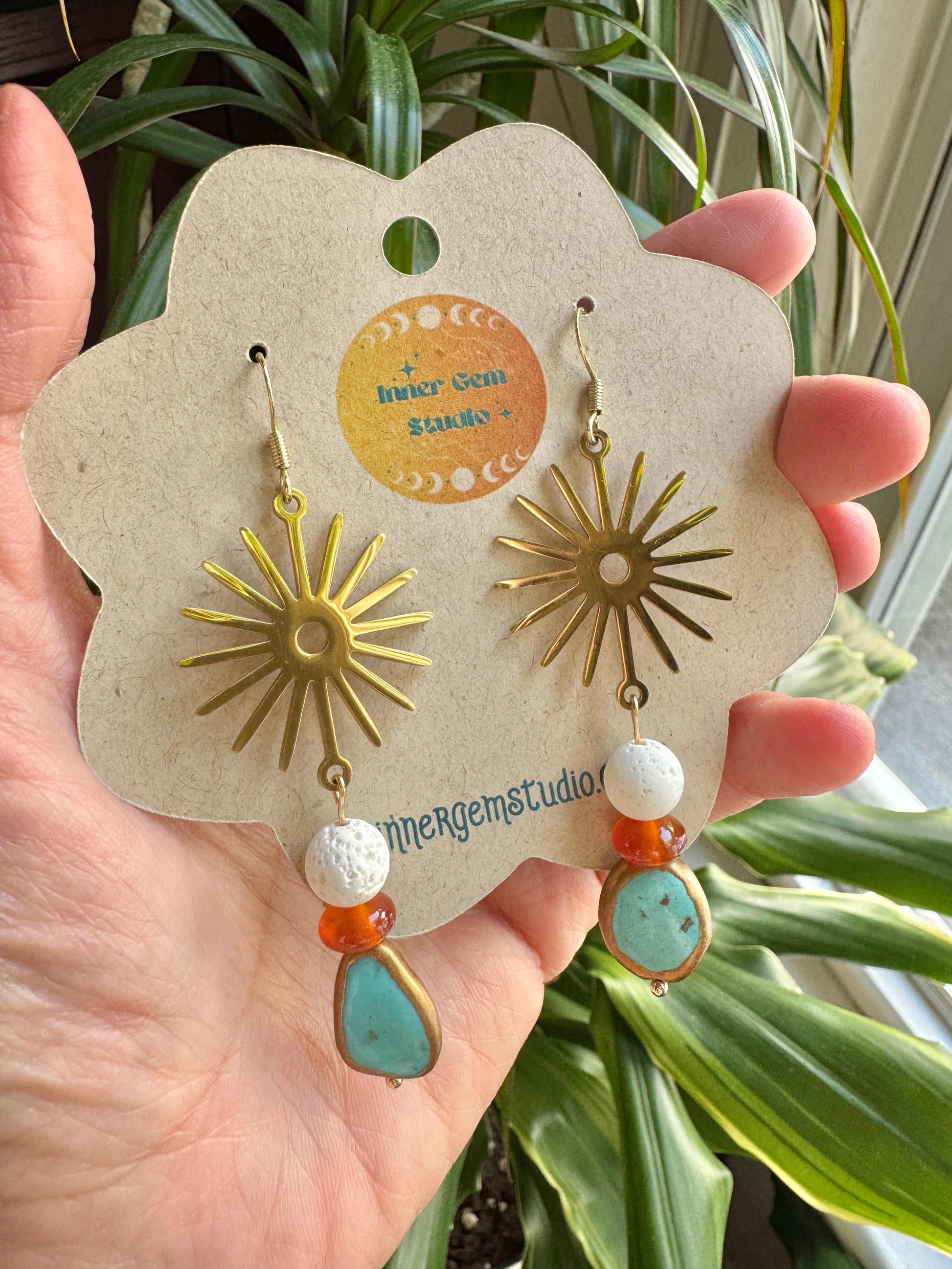 A pair of bohemian-style dangle earrings displayed on a flower-shaped kraft card held in a hand. The earrings feature gold-toned sunburst charms, white textured beads, orange spacer beads, and light blue teardrop beads.