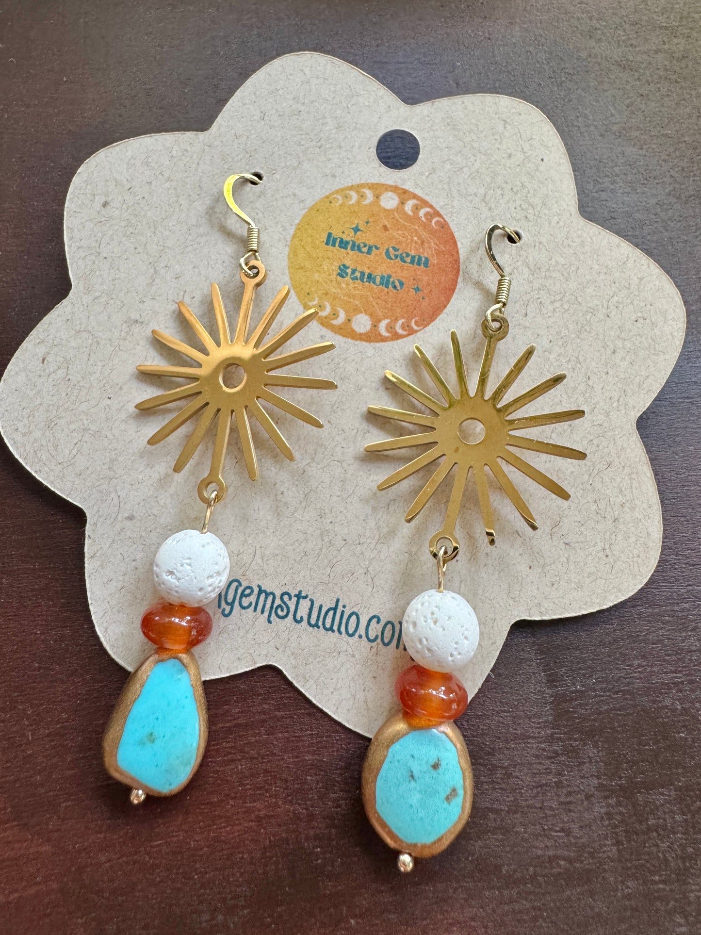 A pair of bohemian-style dangle earrings displayed on a flower-shaped kraft paper card. The earrings feature gold sunburst charms, white textured beads, orange beads, and light blue stone pendants.