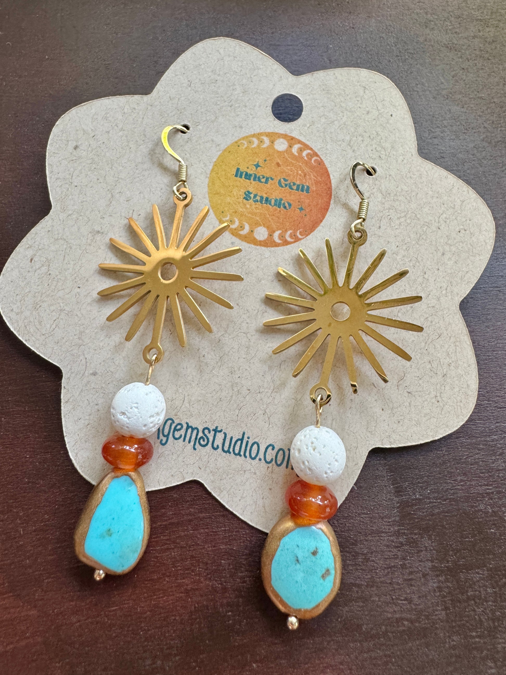 A pair of bohemian-style dangle earrings displayed on a flower-shaped kraft paper card. The earrings feature gold sunburst charms, white textured beads, orange beads, and light blue stone pendants.