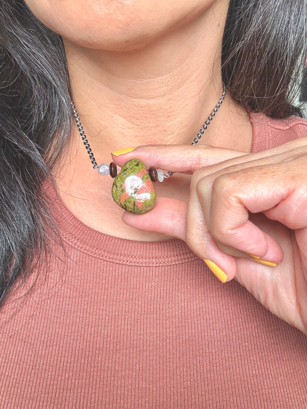 A close-up of a person wearing a silver chain necklace with a small, round, hand-painted green stone pendant featuring pink and white details, held by a hand with yellow nails.