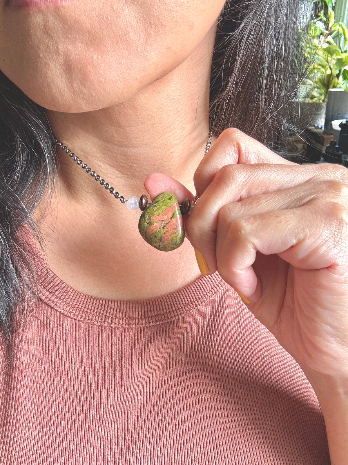 A close-up of a person wearing a silver chain necklace with a polished green stone pendant featuring pinkish-red veining, while holding the pendant near their neck.