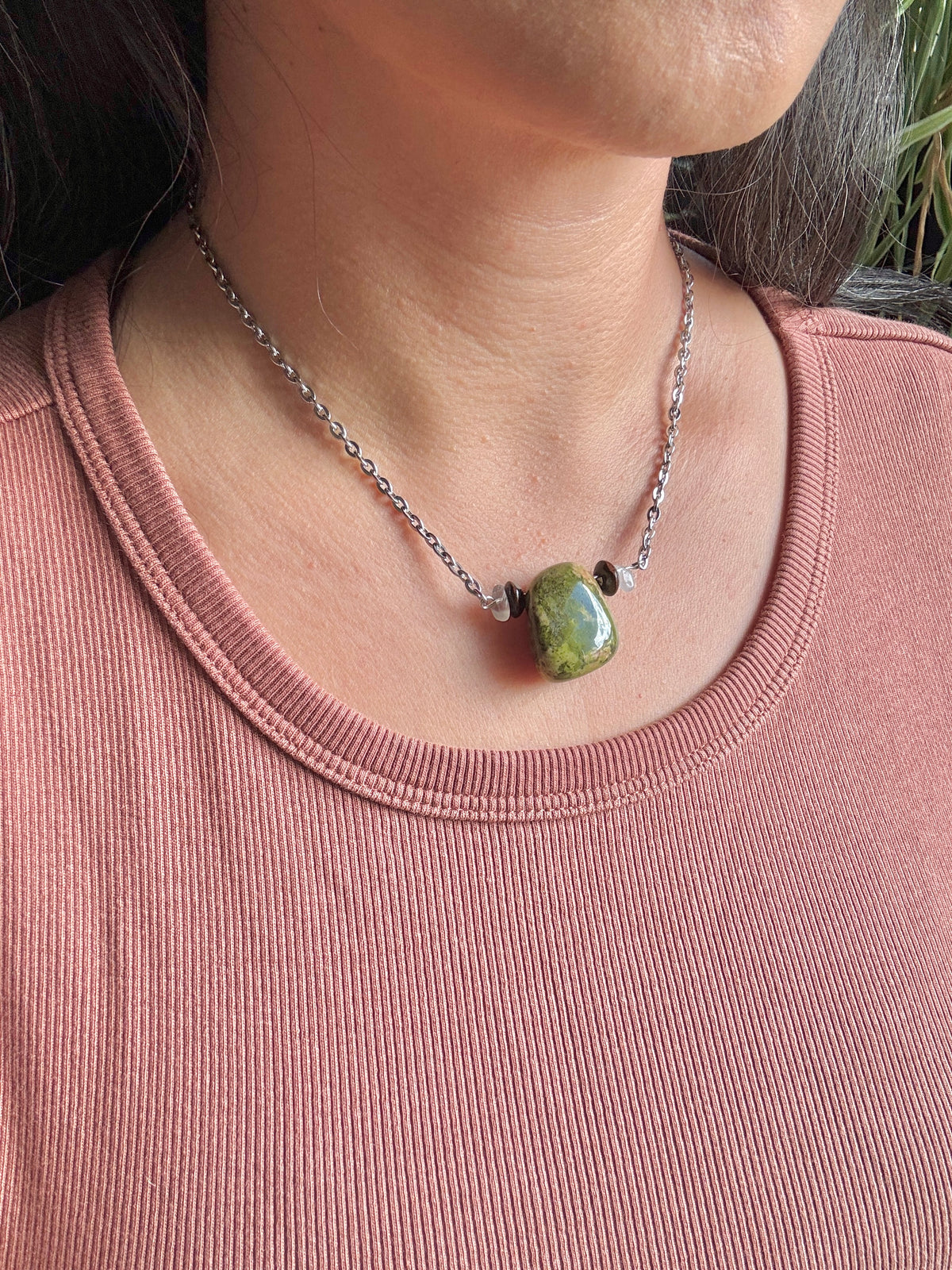 A close-up of a person wearing a silver chain necklace with a polished green stone pendant, paired with a dusty rose ribbed top.