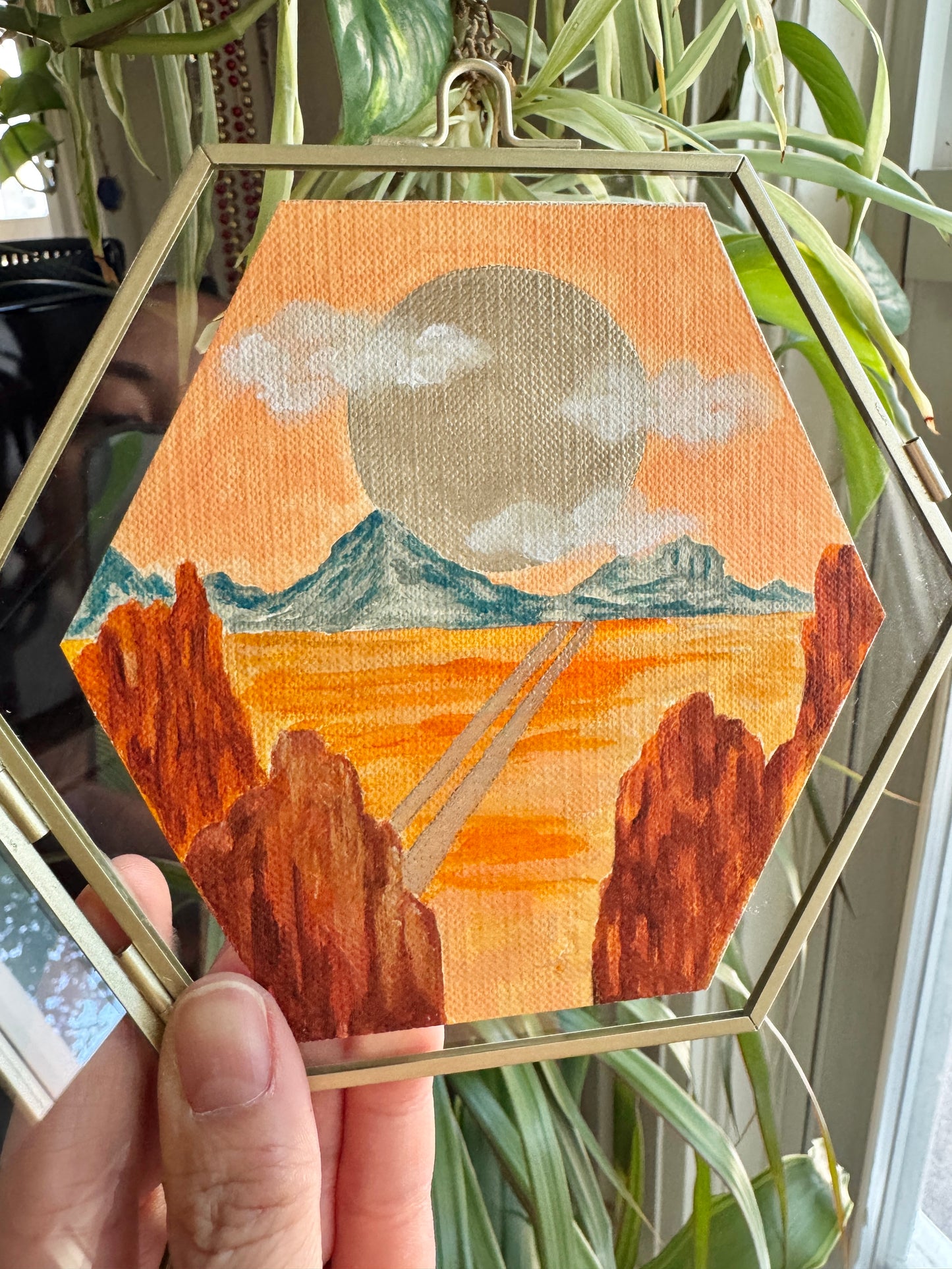 Hand holding up a glass framed desert landscape painting against a plant background
