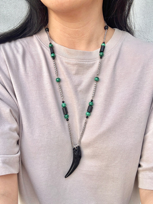 Person wearing a necklace with green beads and a black horn pendant on a plain background