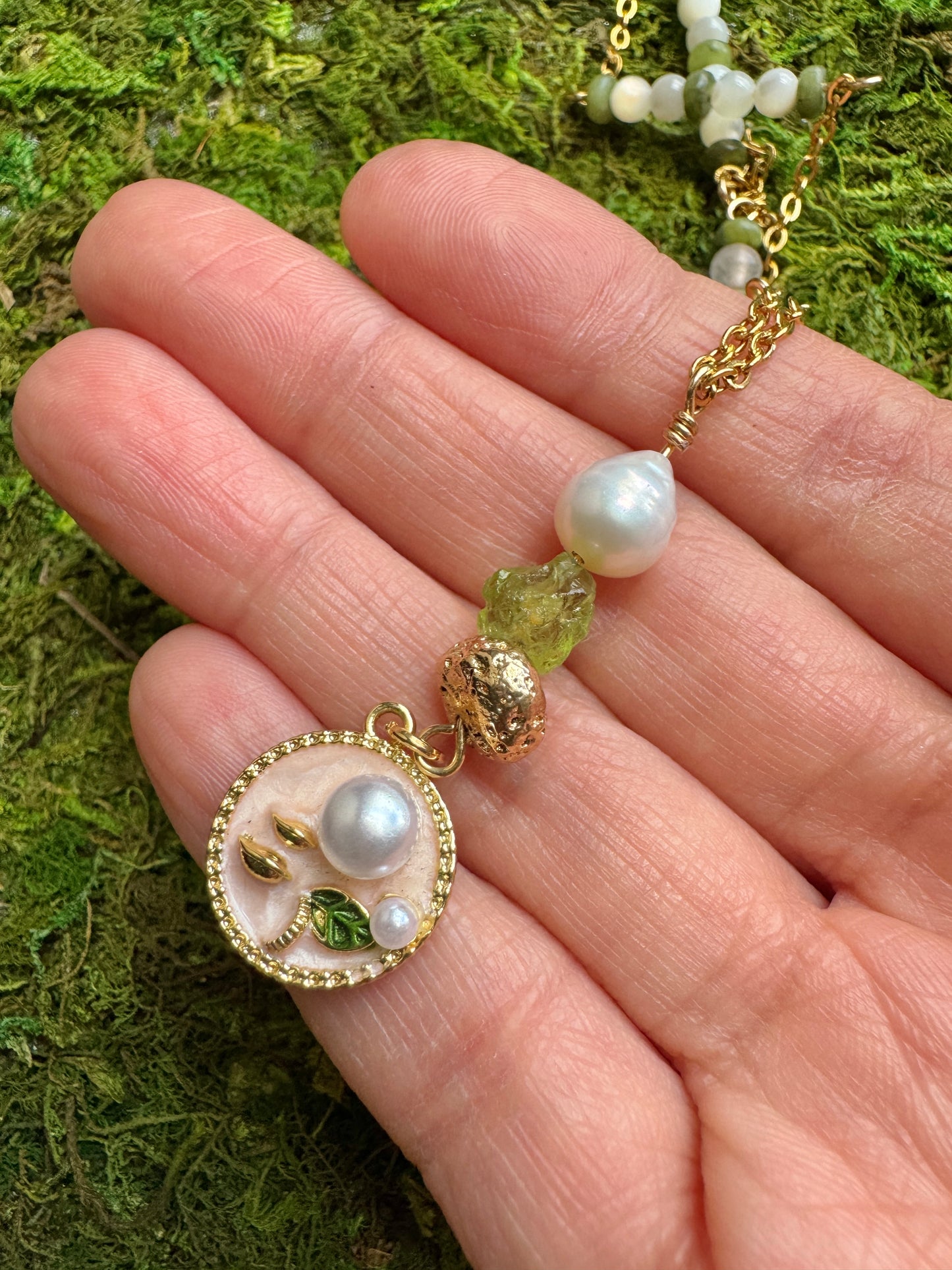 Gold necklace with pearl and green bead pendant held by a hand on a moss background